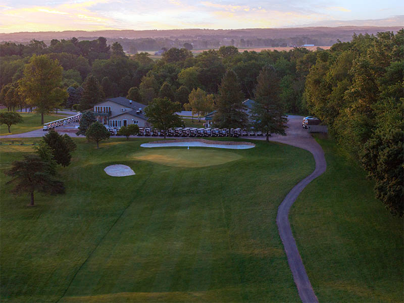 Hole 9 at Flatbush Golf Course in Littlestown, Pennsylvania, featuring a green near the clubhouse, sand bunkers, cart path, golf carts, and sunset countryside views.