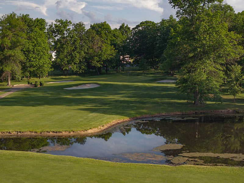 Hole 7 at Flatbush Golf Course in Littlestown, Pennsylvania, featuring a pond in front of the fairway, sand bunkers near the green, and mature trees surrounding the hole.