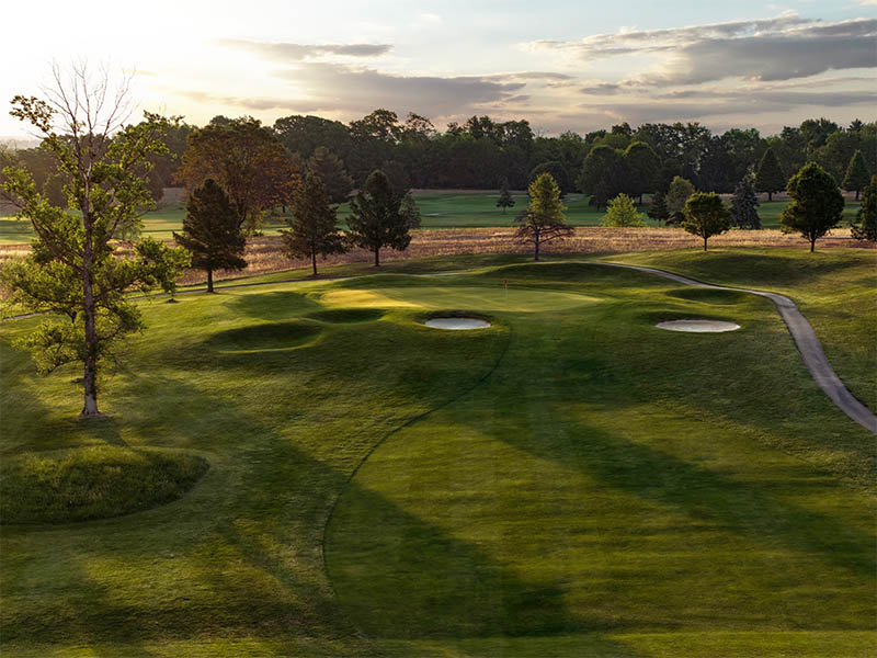 Hole 2 at Flatbush Golf Course in Littlestown, Pennsylvania, featuring an elevated green, sand bunkers, rolling fairway, cart path, and tree-lined sunset views.