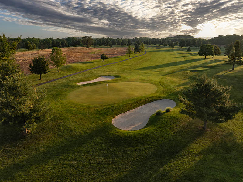 Flatbush Golf Course in Littlestown, Pennsylvania, featuring a green with sand bunkers, rolling fairway, mature trees, cart path, and dramatic evening sky.