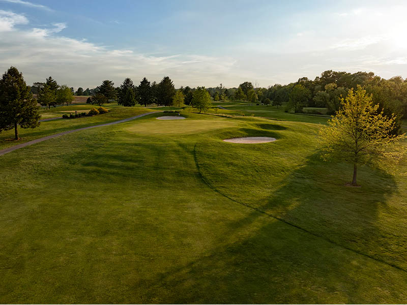 Hole 15 at Flatbush Golf Course in Littlestown, Pennsylvania, featuring a green with two sand bunkers, a tree-lined fairway, cart path, and partly cloudy sky.