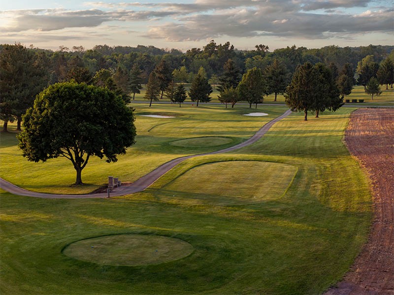 Hole 11 showing an aerial view of the green, two sand bunkers, rolling fairway, cart path, trees, and nearby farmland.