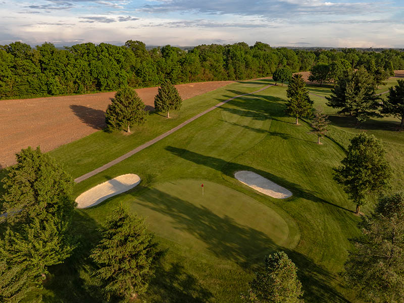 Hole 11 at Flatbush Golf Course in Littlestown, Pennsylvania, showing an aerial view of the green, two sand bunkers, rolling fairway, cart path, trees, and nearby farmland.