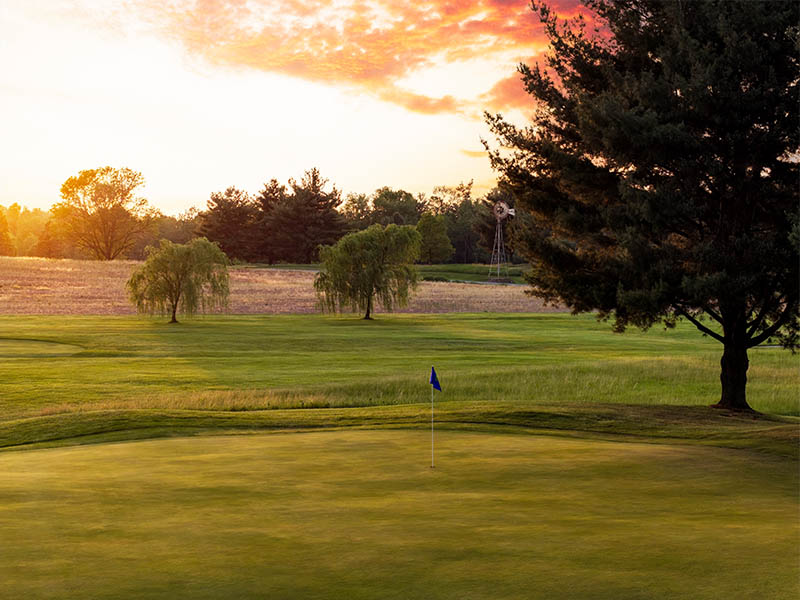 Hole 10 at Flatbush Golf Course in Littlestown, Pennsylvania, featuring a green with a blue flag, open fairway, mature trees, and a colorful sunset sky.